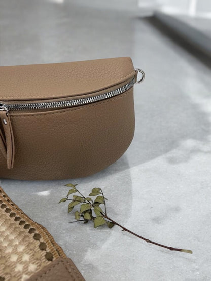 Sac banane simili cuir marron taupe avec fermeture éclair argentée, posé sur surface grise avec branche décorative