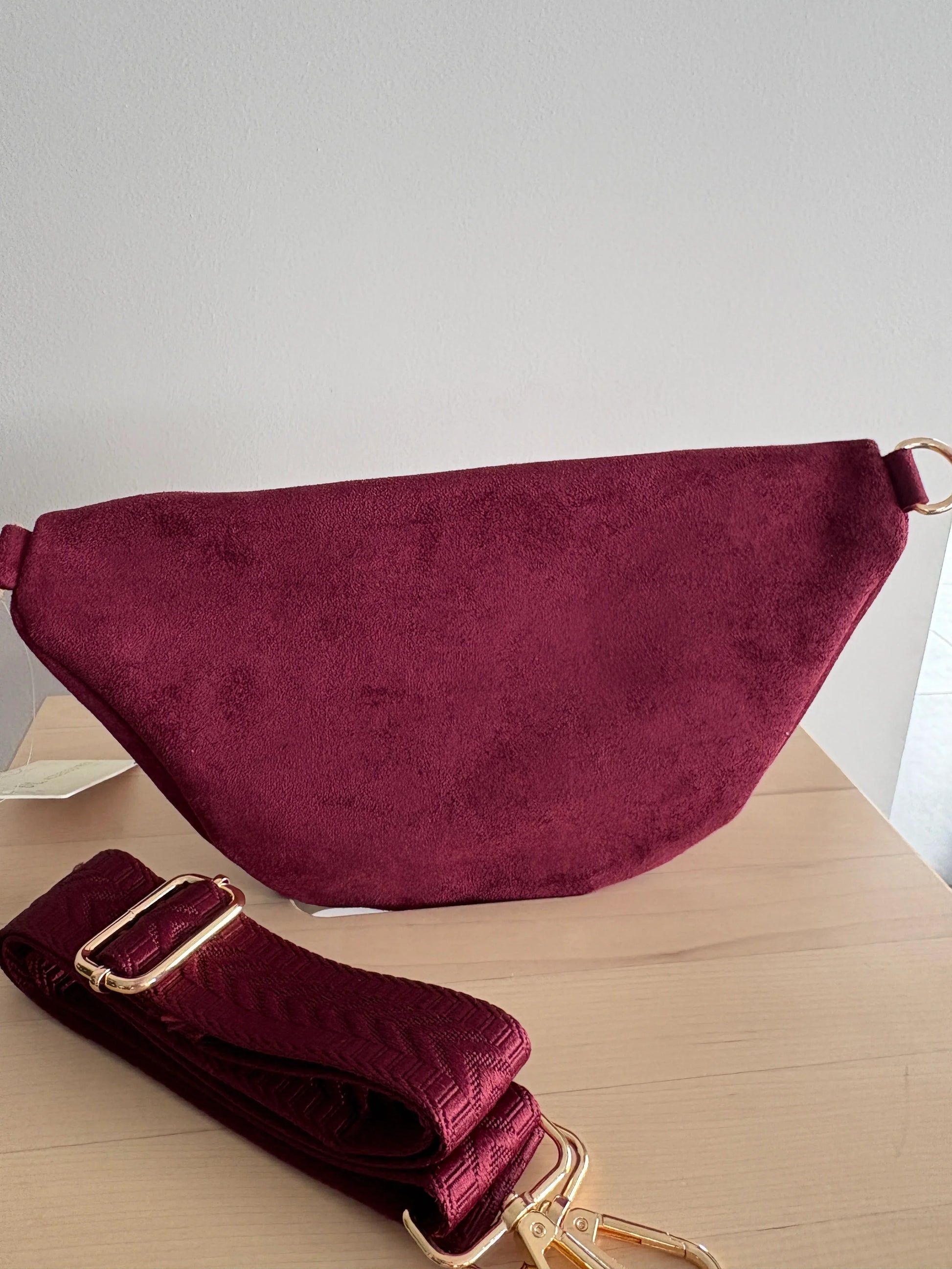Sac banane toucher daim bordeaux avec sangle assortie et fermetures zip dorées, posé sur une table en bois