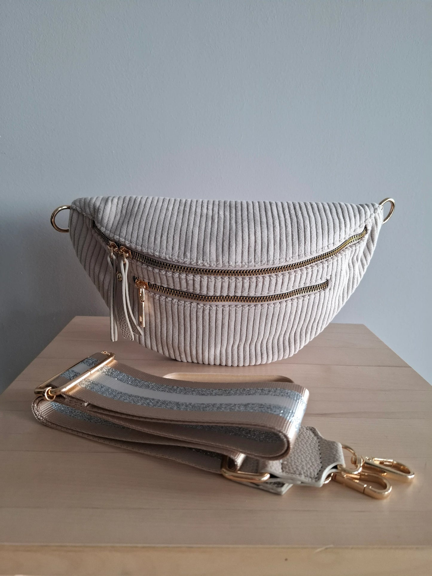 Sac banane velours côtelé beige avec zips dorés et sangle géométrique réglable, posé sur une surface en bois