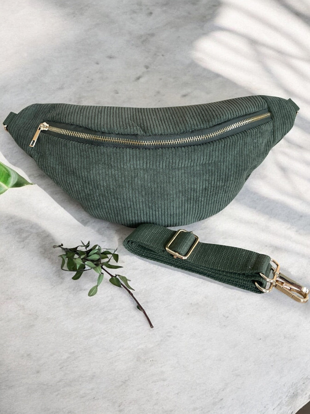 Green corduroy belt bag with gold zipper by Le Petit Placard, displayed on a marble surface