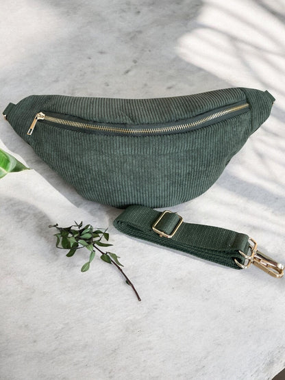 Green corduroy belt bag with gold zipper by Le Petit Placard, displayed on a marble surface