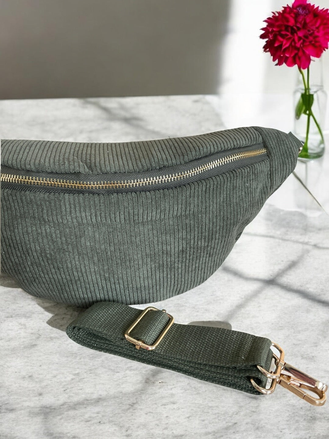 Green corduroy bum bag with gold zipper and strap on marble table, bright pink flower in vase