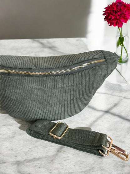 Green corduroy bum bag with gold zipper and strap on marble table, bright pink flower in vase