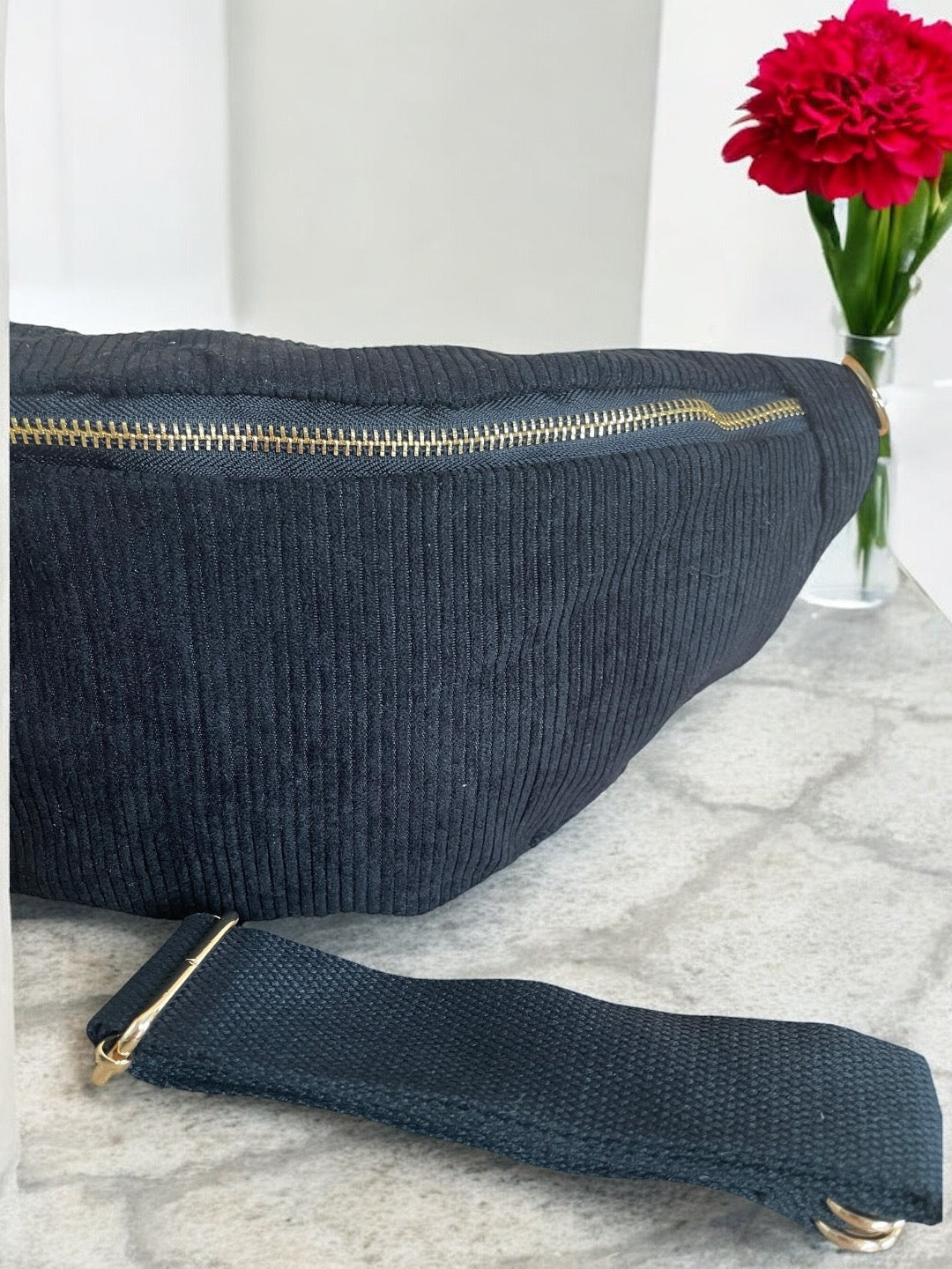 Black textured sac banane with gold zipper on marble surface, red flower in background