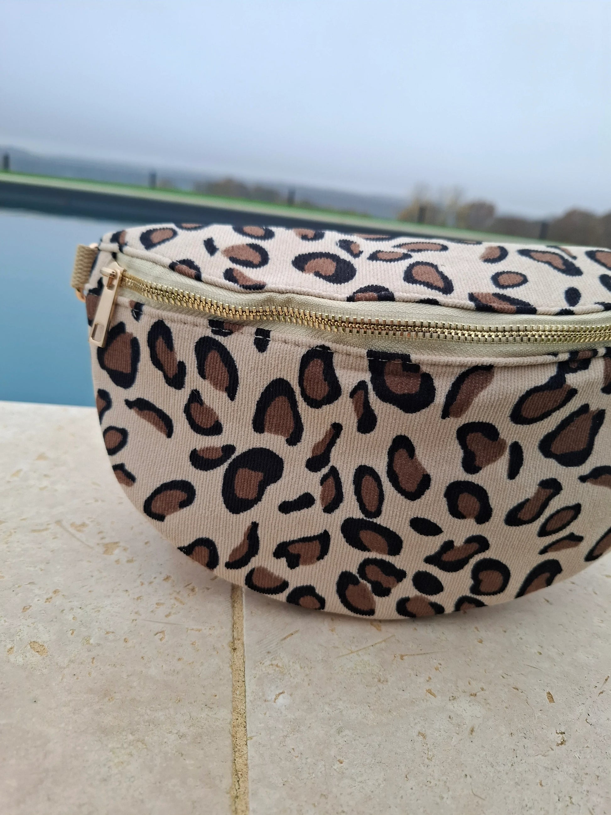 Sac banane velours côtelé imprimé léopard beige et marron avec zip doré, posé sur une margelle de piscine
