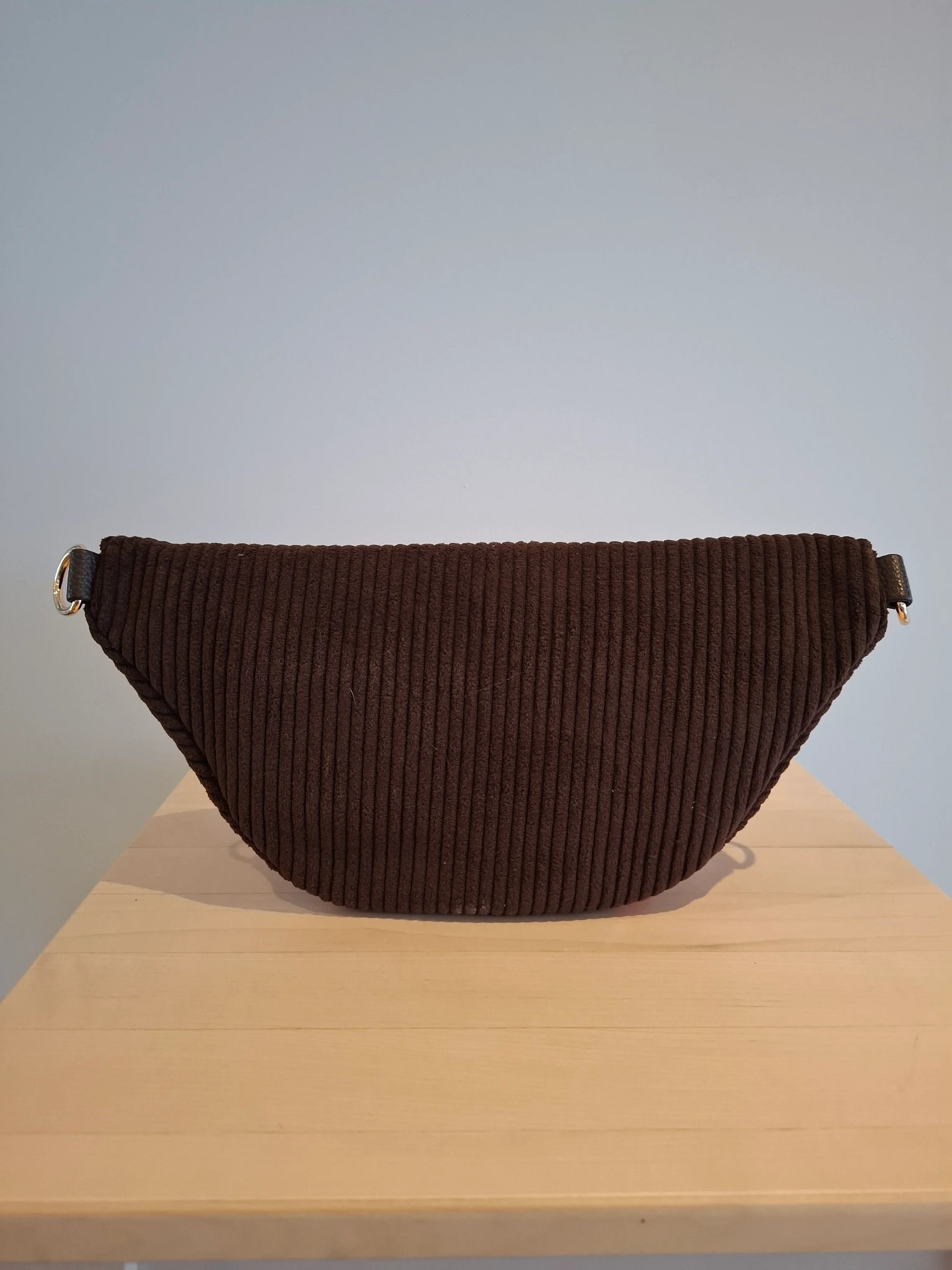 Sac banane velours côtelé marron chocolat foncé avec sangle réglable et anneaux dorés, posé sur une surface en bois
