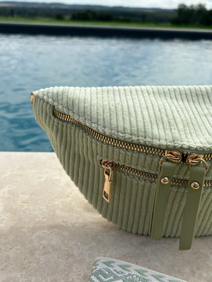 Sac banane velours côtelé vert d'eau avec 2 poches zip dorées et sangle fantaisie, posé au bord d'une piscine