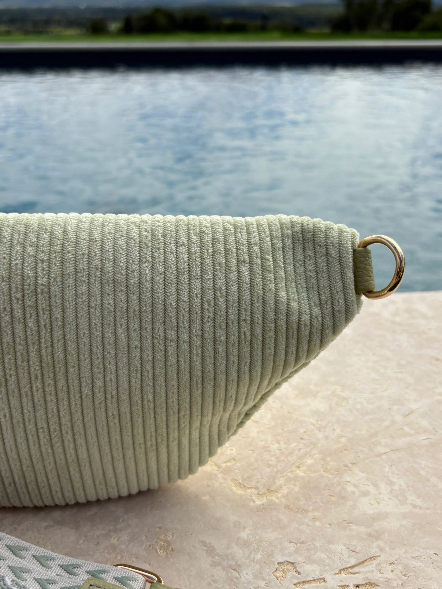 Sac banane velours côtelé vert d'eau avec anneau doré, posé au bord d'une piscine extérieure