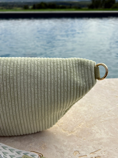 Sac banane velours côtelé vert d'eau avec anneau doré, posé au bord d'une piscine extérieure