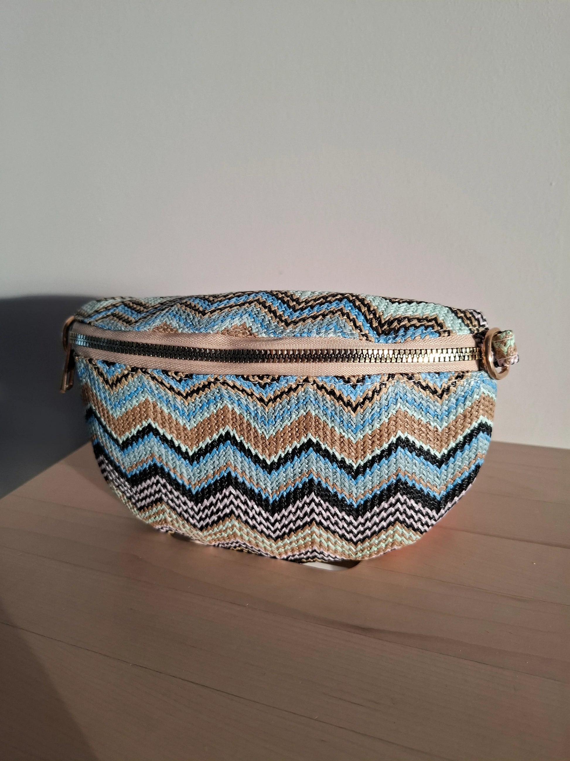 Sac banane zigzag multicolore bleu beige noir avec fermeture éclair et bandoulière dorée, posé sur une table