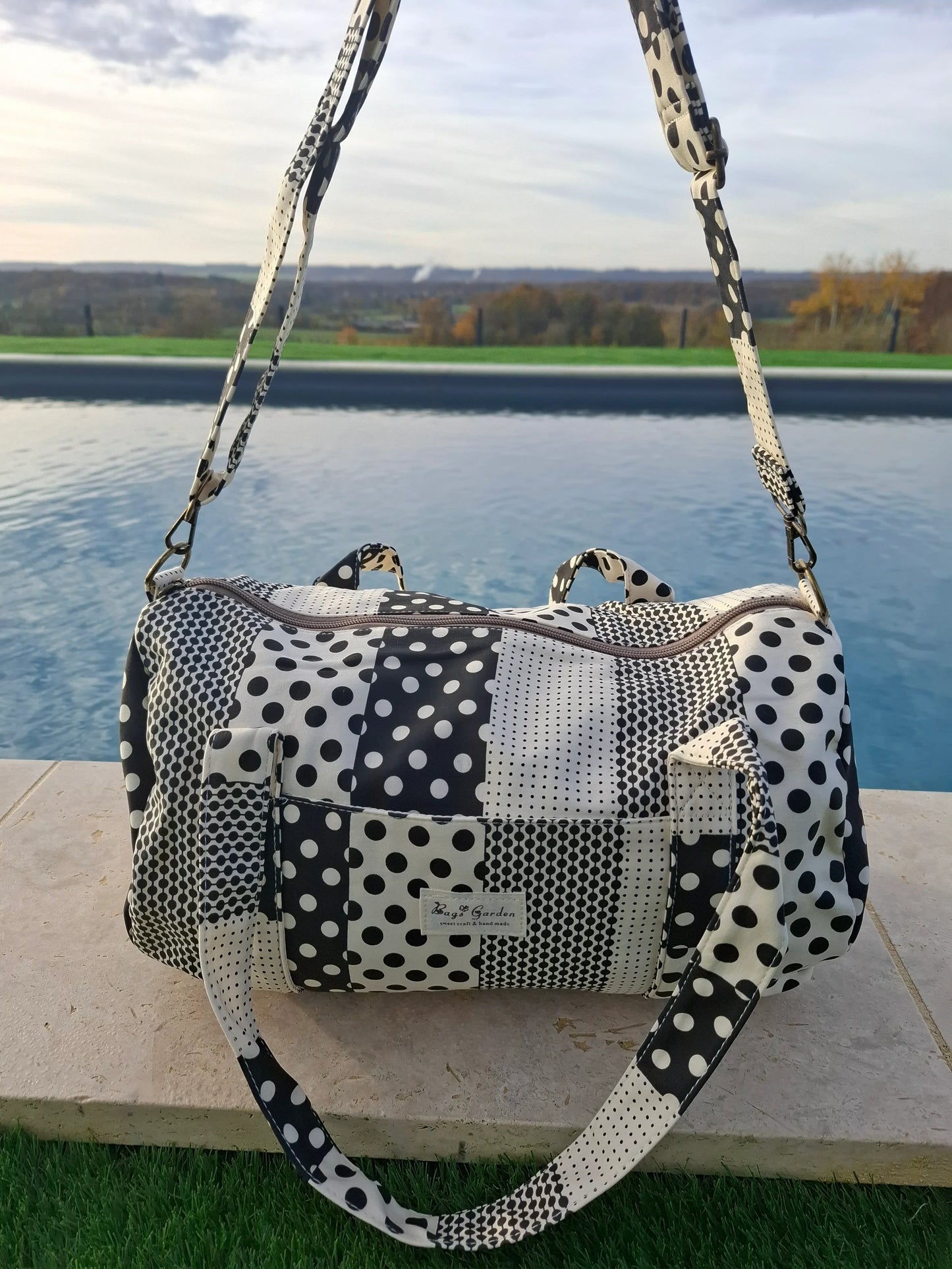Sac polochon tissu coton patchwork pois noir et blanc avec bandoulière, posé au bord d'une piscine
