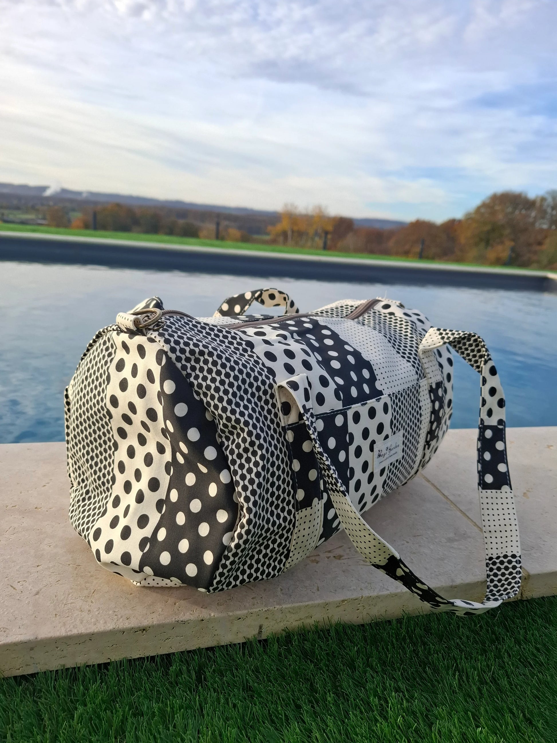 Sac polochon tissu coton patchwork pois noir et blanc posé au bord d'une piscine extérieure