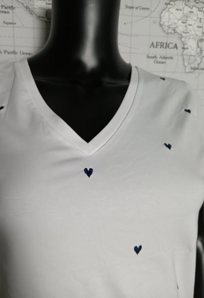 T-shirt blanc à col V avec petits cœurs bleus brodés, présenté sur un mannequin. Le motif reste discret et élégant.