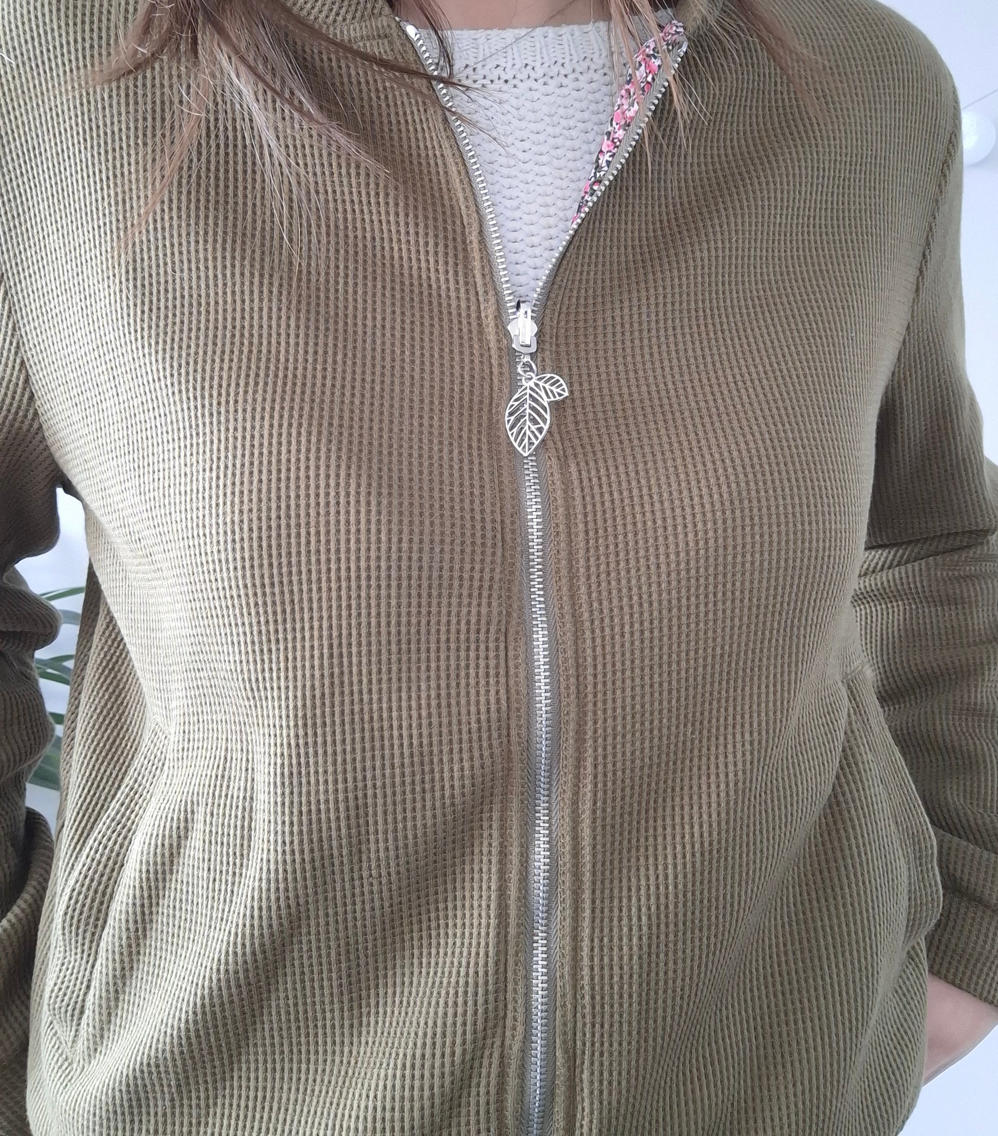 Close-up of a khaki textured zip-up jacket with leaf zipper pull, worn over a white sweater
