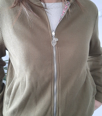 Close-up of a khaki textured zip-up jacket with leaf zipper pull, worn over a white sweater