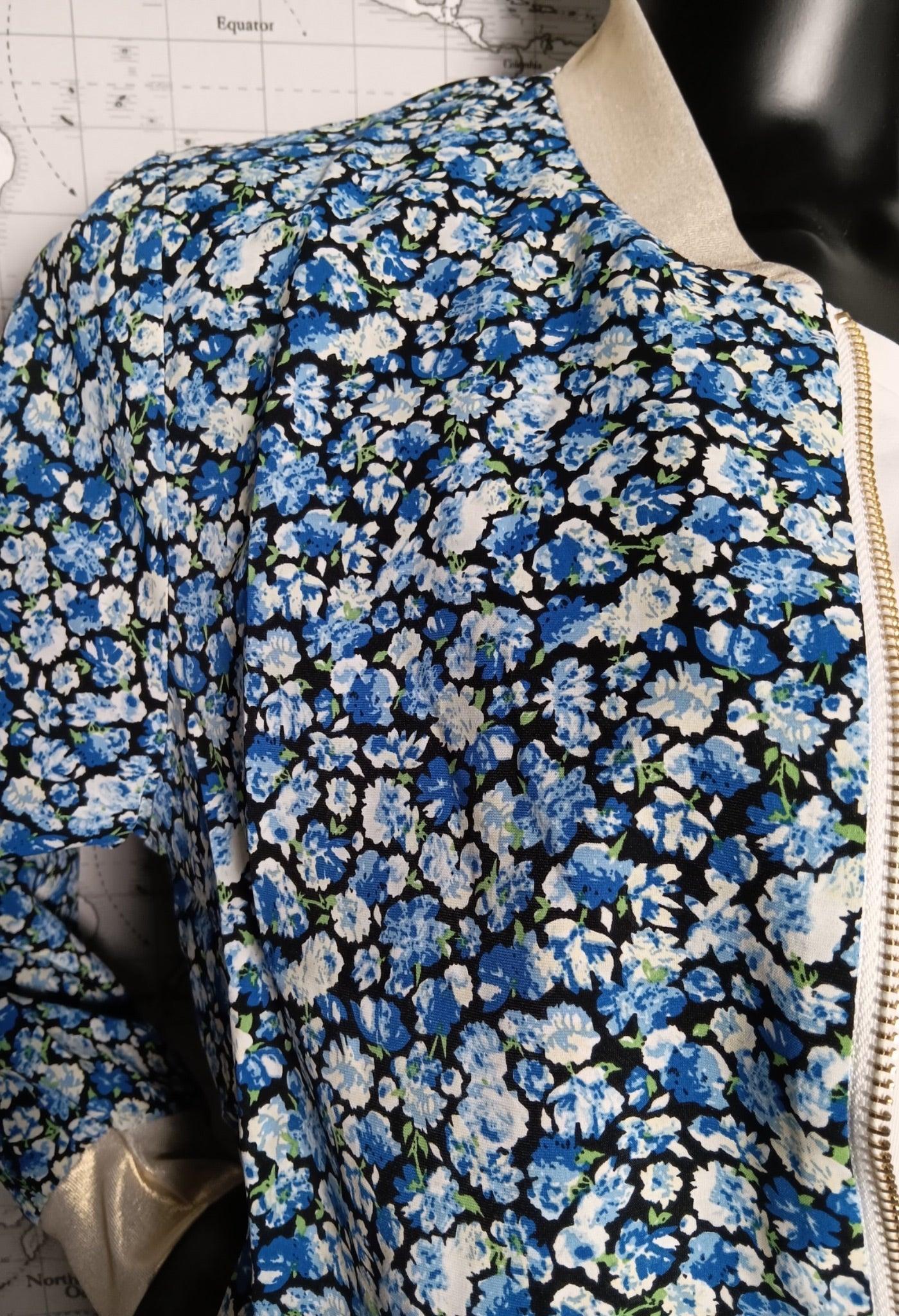Veste femme imprimé fleurs bleues et blancs sur fond noir, avec fermeture éclair dorée visible.
