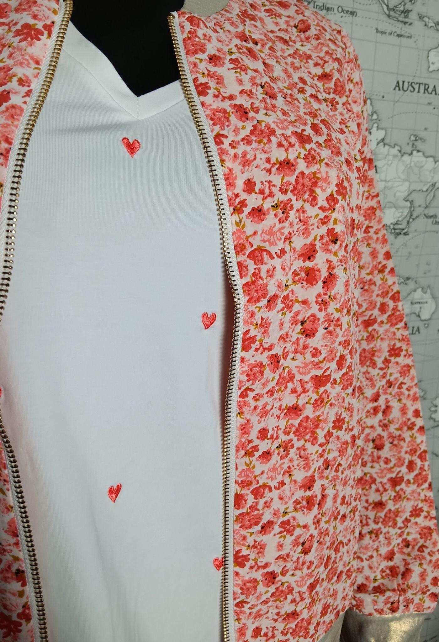 Veste bomber fleurie rouge et orangé légèrement ouverte sur un tee-shirt blanc à petits cœurs rouges, zippée doré.