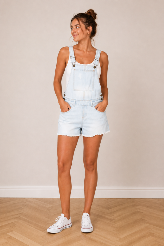 Salopette short en jean délavé, style vintage estival, femme debout portant short overalls bleus clair avec baskets blancs.