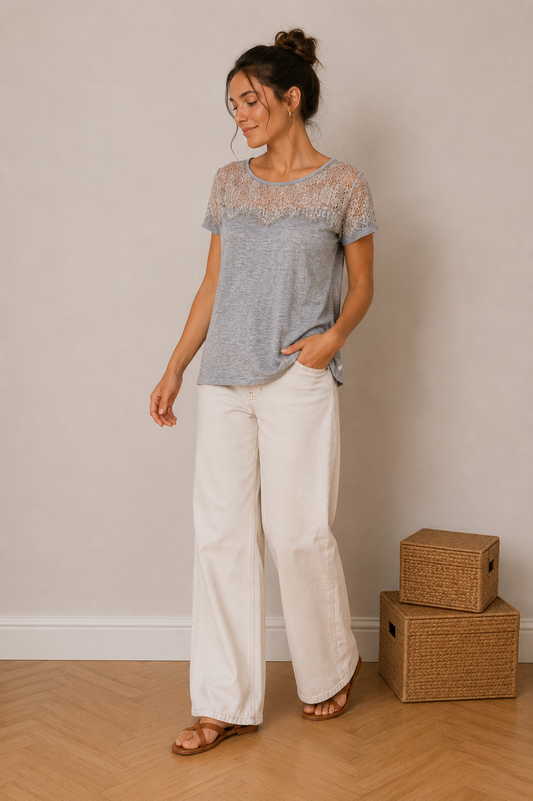 T-shirt gris chiné à empiècement dentelle florale sur le haut, femme pose décontractée, pantalons blancs ample, sandales, intérieur neutre.