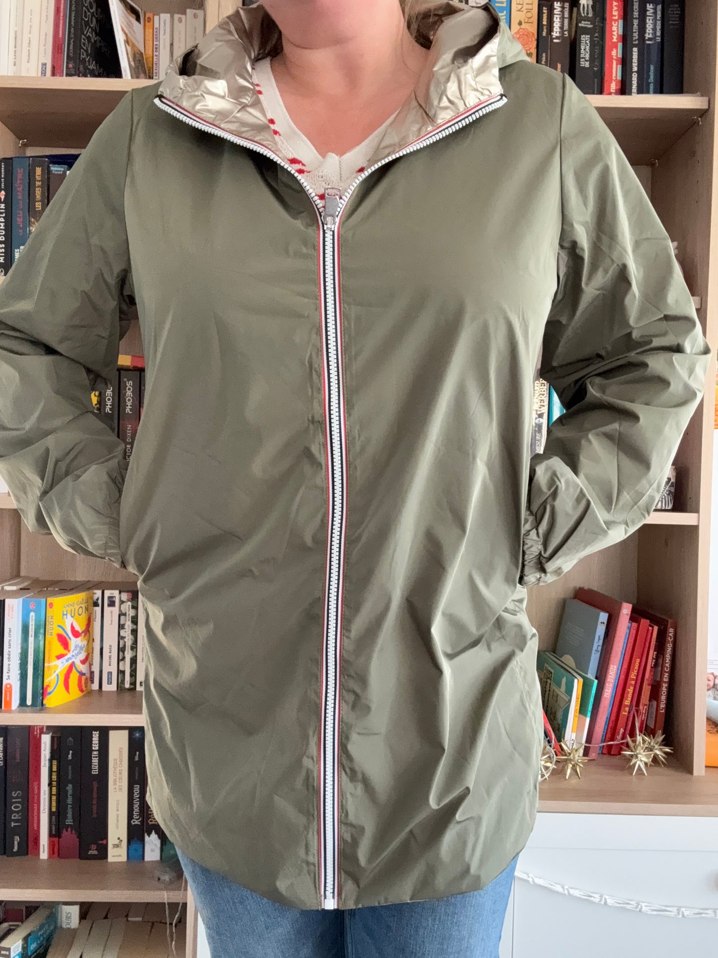 Imperméable kaki femme Melya Melody avec doublure dorée, fermeture éclair blanche et rouge, porté devant une bibliothèque