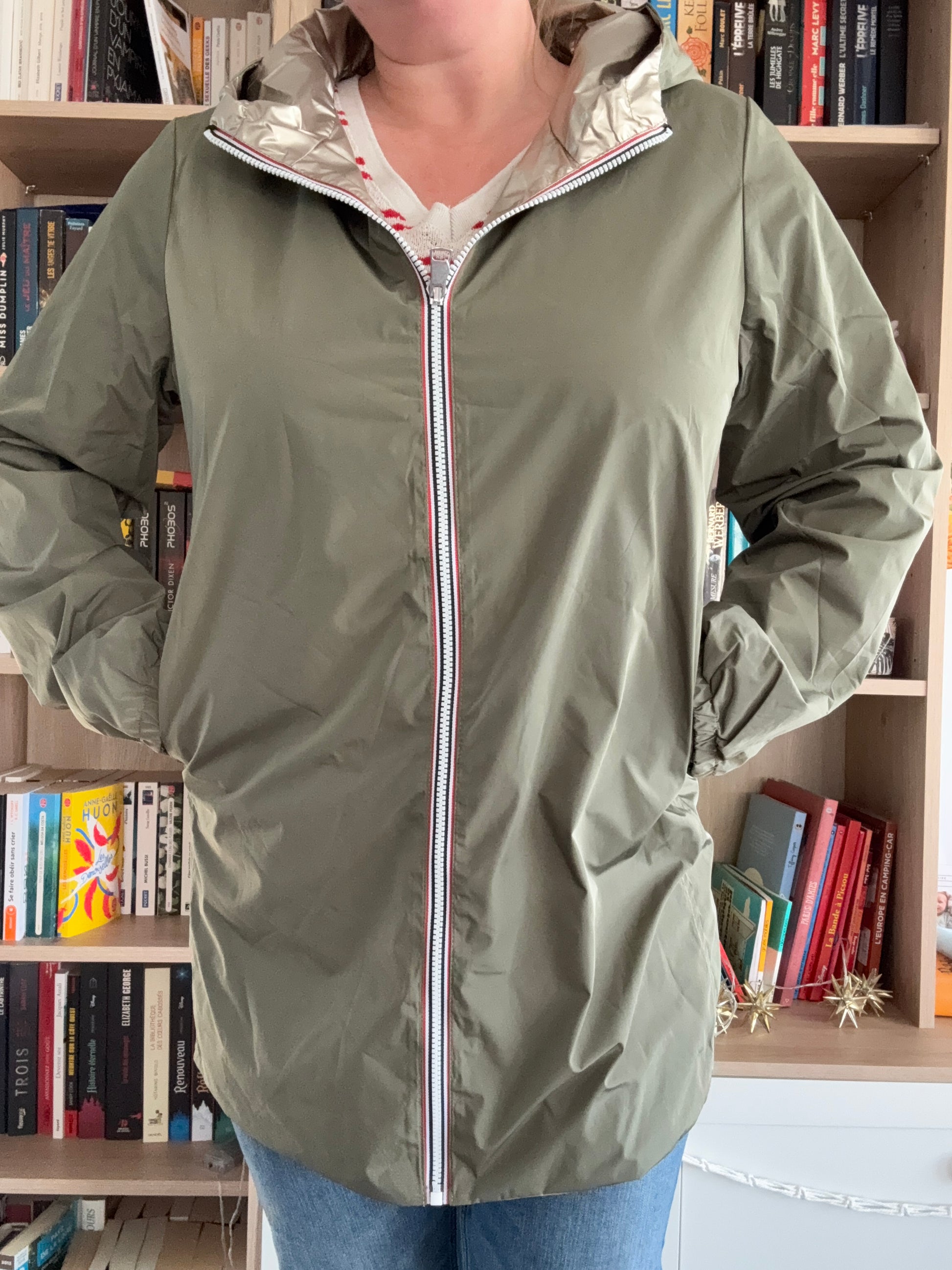 Imperméable kaki femme Melya Melody avec doublure dorée, fermeture éclair blanche et rouge, porté devant une bibliothèque