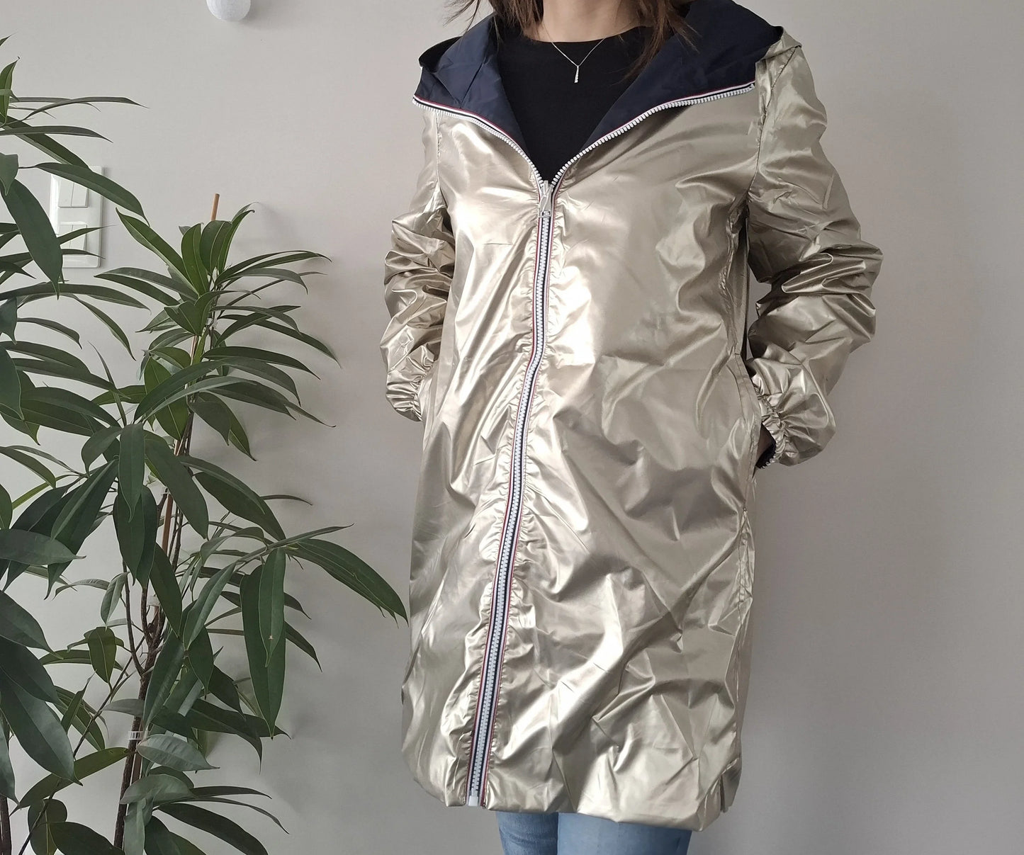 Woman wearing a shiny gold Laura Jo raincoat from Le Petit Placard, standing indoors by a plant.