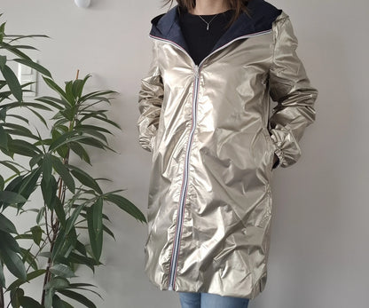 Woman wearing a shiny gold Laura Jo raincoat from Le Petit Placard, standing indoors by a plant.