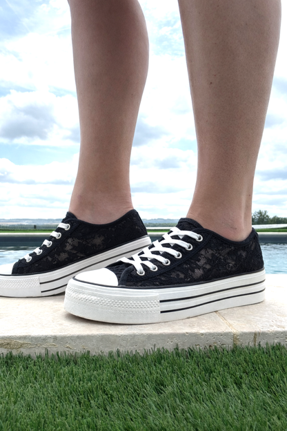 Basket Plateforme Dentelle Noire Femme avec semelle épaisse: pieds portant des tennis noires en dentelle, lacets blancs, grande semelle blanche à rayures noires.